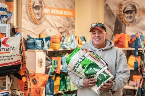 Der Besitzer von BEAD steht mit einer recycelten Tasche im Arm in seinem Laden. Im Hintergrund stehen Regale mit verschiedenen Taschen. 