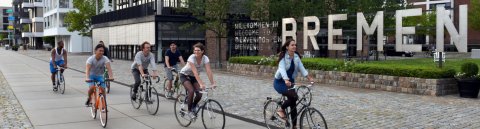 Junge Menschen auf fahren Fahrrad. Hinter ihnen Häuser und der Schriftzug "Willkommen in Bremen"