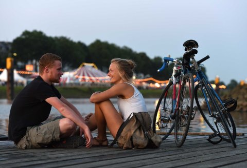 Ein junger Mann und eine junge Frau sitzen zur Abenddämmerung in sommerlicher Kleidung neben ihren Rädern auf einem Steg an der Weser, im Hintergrund die Lichter der Breminale.