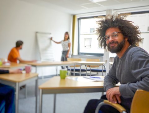 Ein Mann mit wuschelig-lockigem Haar und einer Brille sitz in einem Kursraum und lächelt in die Kamera. Im Hintergrund andere Kursteilnehmende und eine Lehrerin.