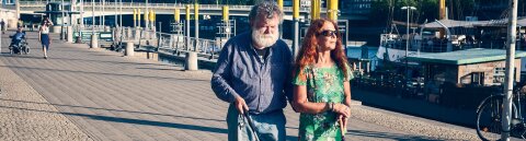 Ein Mann und eine Frau, beide mit Blindenstock, gehen an einem Fluss spazieren. Im Hintergrund sind Schiffe und eine Brücke zu sehen.