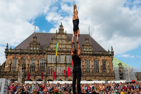 Zwei Akrobat*innen führen auf dem Bremer Marktplatz ein Kunststück auf. Im Hintergrund ist das Rathaus und Publikum zu sehen. Der Himmel ist blau mit ein paar Wolken.