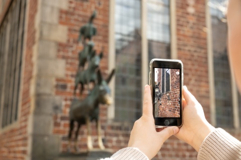 Ein Smartphone mit animierten Stadtmusikanten Figuren wird vor die Statue der Stadtmusikanten gehalten.