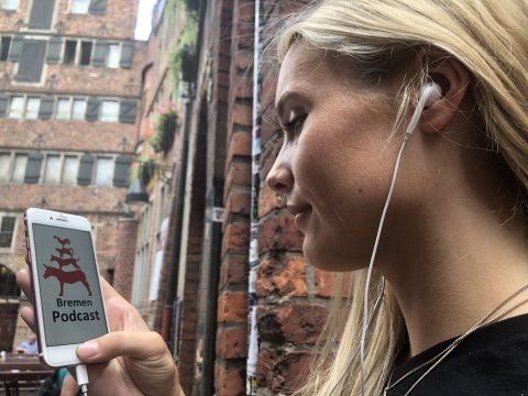 Eine junge Frau hält ihr Smartphone in der Hand. Auf dem Bildschirm ist das Logo des Bremen-Podcasts zu sehen.