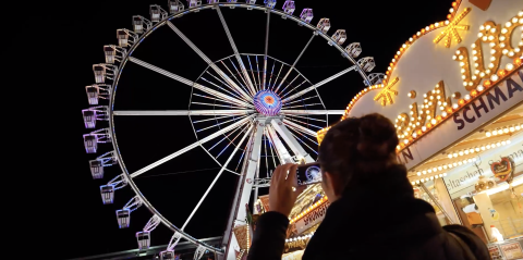 Eine Frau macht mit ihrem Smartphone ein Foto von einem beleuchteten Riesenrad auf dem Jahrmarkt.