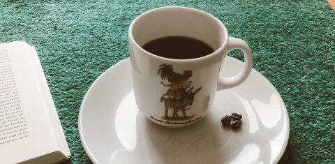 Eine weiße Kaffeetasse, auf der die Bremer Stadtmusikanten abgebildet sind, steht auf einem weißen Unterteller. Beide stehen auf einem türkisfarbenen Teppich. Links ragt noch ein Teil eines Buches ins Bild.