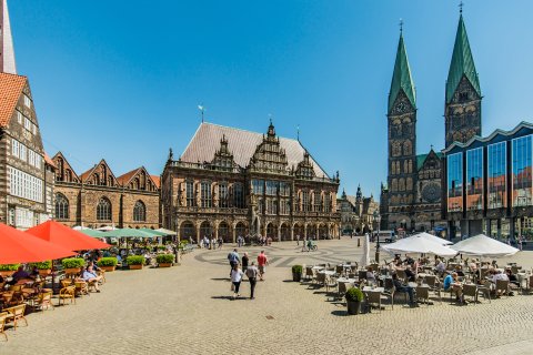 Aufnahme eines Platzes, auf dem Tische, Stühle und Sonnenschirme stehen. Der Platz ist von mehreren Gebäuden umgeben. 