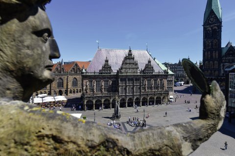 Blick auf ein steinernes, historisches Gebäude, welches auf einem Platz steht, auf dem sich mehrere Menschen aufhalten. Zudem zeigt die Aufnahme einen Turm des Bremer Doms. 