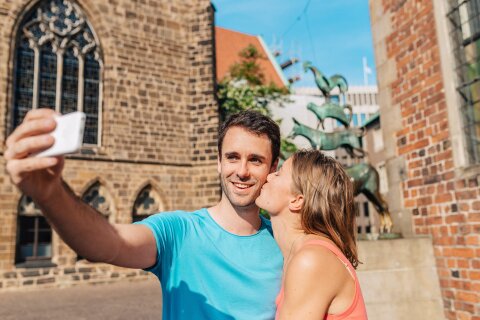 Eine Frau küsst einen Mann auf die Wange. Der Mann hält ein Handy in der Hand und schießt ein Foto von ihnen vor den Bremer Stadtmusikanten.