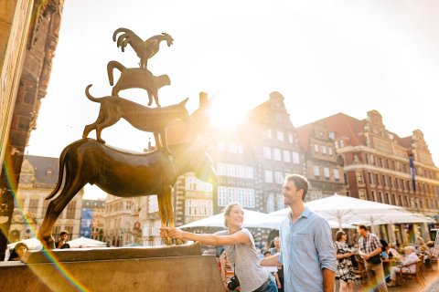 Eine Frau umfasst die Füße des Esels der Bremer Stadtmusikanten. Sie lächelt dabei einen Mann an. 