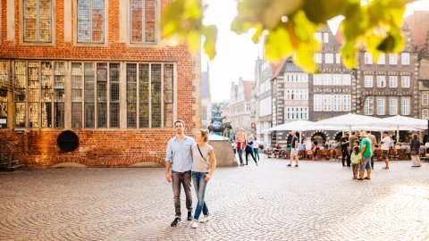 Eine Frau und ein Mann laufen am Bremer Rathaus vorbei. Im Hintergrund sieht man mehrere Menschen bei den Bremer Stadtmusikanten stehen. 