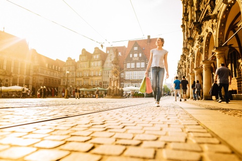 A woman walks across a square with shopping bags. Historical buildings can be seen in the background. The sun is shining.