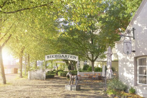Eine sommerliche Aufnahme zeigt einen kleinen Platz gesäumt von grünen Bäumen mit Eingang zu einem Biergarten.