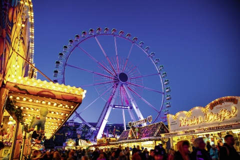 Die Osterwiese am Abend. Menschen schlendern an den Buden vorbei. Im Hintergrund leuchtet ein Riesenrad