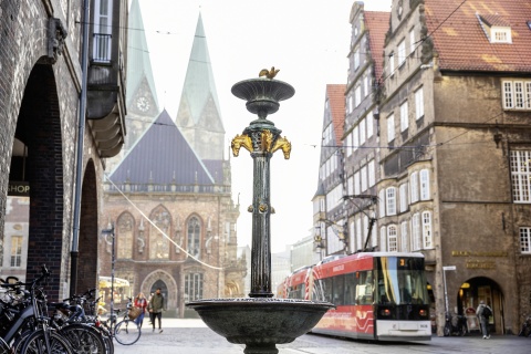 Ein goldverzierter Brunnen in einer Bremer Einkaufsstraße. Im Hintergrund ist der Bremer Dom und eine Straßenbahn zu sehen.