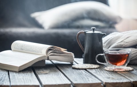 Ein aufgeschlagenes Buch liegt auf einem Holztisch, daneben steht eine Teekanne und ein gefülltes Teeglas. Im Hintergrund ein Sofa mit Kissen.