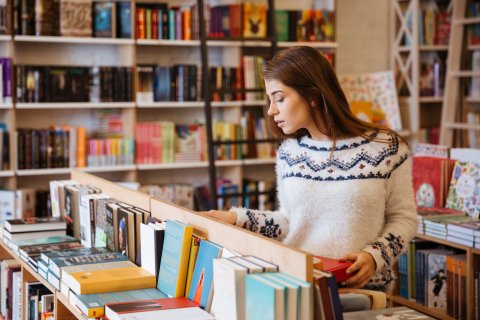 A young woman is standing in front of a table-high shelf in a bookshop, looking at the books. In the background are large bookshelves.