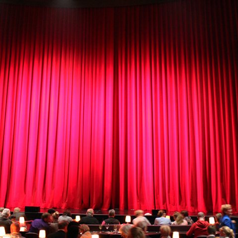 Ein großer roter geschlossener Vorhang, vor dem einige Menschen sitzen.