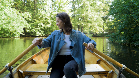 Eine Frau sitzt in einem Ruderboot und hält ein Ruder in jeder Hand. Sie schaut lachend zur Seite. Das Ruderboot befindet sich auf dem Wasser im Bremer Bürgerpark und ist von grünen Bäumen umgeben. Im Hintergrund ist eine Brücke zu sehen.