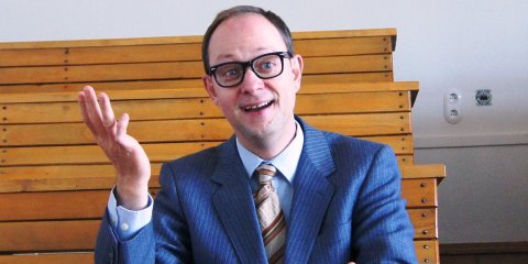 Christian Schliehe sitzt im Anzug und hebt die Hand fragend in die Luft, während er lächelt.