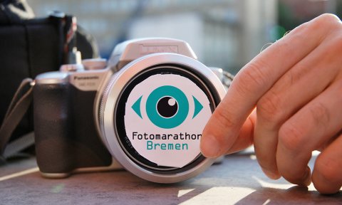 Eine Hand hält das Logo des Fotomarathons vor ein Kameraobjektiv; Quelle: Fotomarathon Bremen/Kerstin Graf