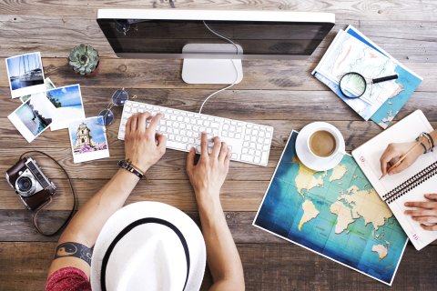 Ein Mann plant am Computer eine Reise. Neben ihm liegen eine Karte und Bilder.