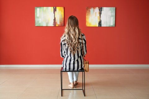 Eine Frau sitzt mit dem Rücken zur Kamera vor einer Wand, an der zwei Bilder aufgehängt sind