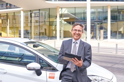 Mann im Anzug steht, ein Tablet in der Hand, grinsend vor einem Auto.