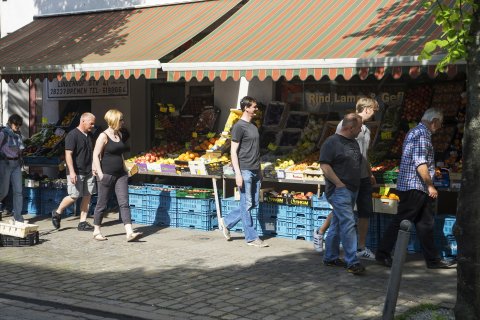 Menschen schlendern vor einem Lebensmittelgeschäft in der Lindenhofstraße. In der Auslage sind viele Obst- und Gemüsesorten zu sehen. (Quelle: Kultur vor Ort e.V.)
