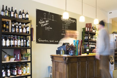 Der Weinladen GluckGluck von innen. Links steht ein Regal mit verschiedenen Weinen. In der Mitte steht ein Kassentresen. Ein Mann und eine Frau stehen am Kassentresen. 