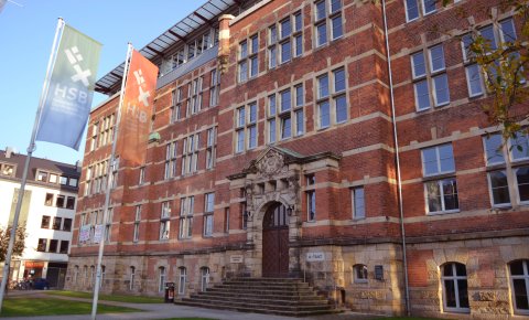 Außenansicht eines Backsteingebäudes, im Vordergrund wehen zwei Fahnen mit der Aufschrift 'Hochschule Bremen'; Quelle: WFB/bremen.online - MDR