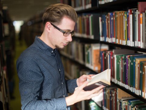 Man sieht einen Mann mit Brille. Er liest ein Buch. Hinter ihm sieht man volle Bücherregale.