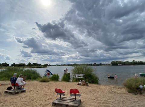 Ein Sandstrand direkt am Hemelinger See. Stühle stehen auf Paletten und mehrere Menschen baden im Wasser.
