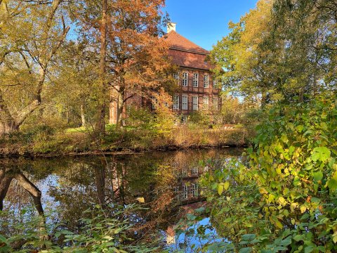 Das von herbstlichen Bäumen und Büschen sowie einem Gewässer umgebene Schloss Schönebeck an einem sonnigen Herbsttag.