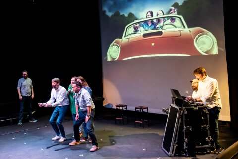 Eine Improvisationsgruppe von Männern und Frauen steht in einem Theater auf einer Bühne. Im Hintergrund wird ein Bild von ihnen in einem Auto an eine Wand projiziert. 