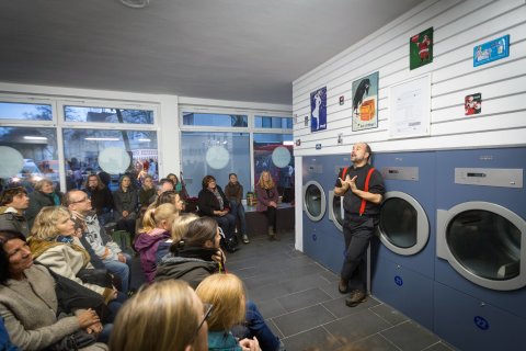 Ein Mann erzählt vor Publikum in einem Waschsalon.