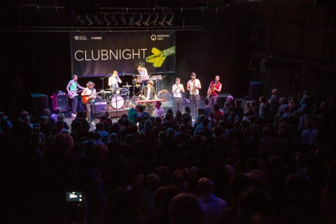Ein Konzert während der jazzahead Clubnight. Eine Band steht hell erleuchtet auf der Bühne, vor dem vollen dunklen Publikum.