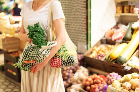 Eine Person hat zwei grob gewebte Einkaufstaschen mit Obst und Gemüse bei sich. Einen hat sie auf der Schulter liegen und den anderen in der Hand. Im Hintergrund befindet sich weiteres Obst und Gemüse.
