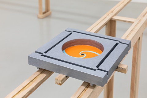 Auf einer Holzkonstruktion ist ein graues Quadrat montiert. In der Mitte ist ein orangener Kreisel.