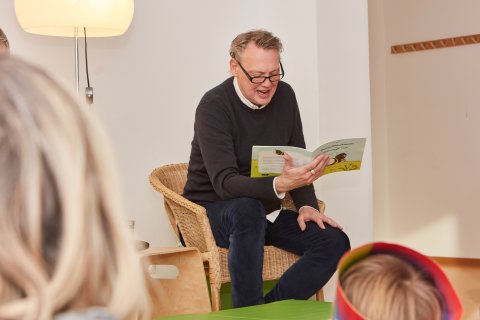 SOS-Kinderdorf Botschafter Dirk Böhling sitzt auf einem Stuhl und liest Kindern ein Buch vor.
