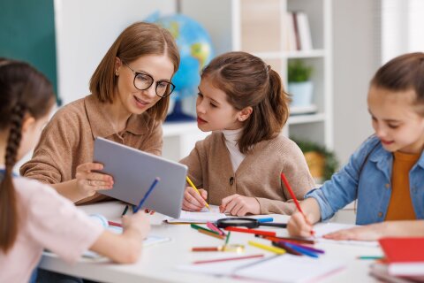 Drei Schülerinnen und eine Lehrerin sitzen an einem Tisch in einem Klassenraum. Die Mädchen malen mit Buntstiften. Eines der Mädchen schaut interessiert auf ein Tablet, das die Lehrerin in der Hand hält.
