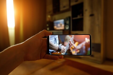 Ein Smartphones wird von einer Hand ins Bild gehalten. Auf dem Bildschirm sind zwei Personen zu sehen, die Gitarre spielen. Im Hintergrund befindet sich ein Wohnzimmer.