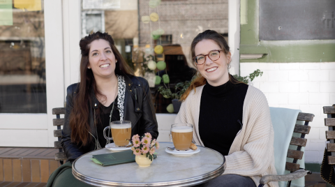 Zwei junge Frauen sitzen vor einem Caféan einem Tisch mit zwei Getränken.