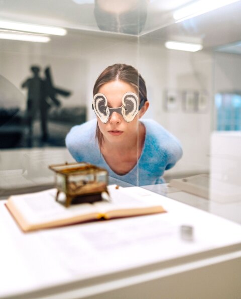 Eine Frau mit einer aus Papier gebastelten Ohren-Brille schaut auf ein Kunst Objekt, das in einem Glaskasten ausgestellt ist. 