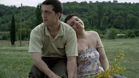 Ein Mann und eine Frau sitzen auf einer Wiese. Die Frau schmiegt sich verliebt an den Mann. Es ist Sommer. Im Hintergrund ist eine hügelige Landschaft mit Bäumen.