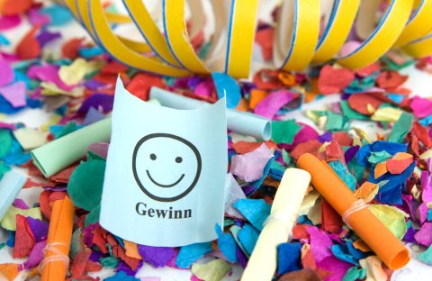 Viele bunte Lose und Luftschlangen liegen auf einem weißen Tisch. Ein Los ist geöffnet, darauf steht Gewinn mit einem lächelnden Smliley.