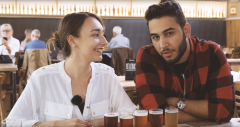 Eine junge Frau und ein junger Mann sitzen an einem Tisch. Vor ihnen steht eine Reihe mit kleinen, gefüllten Biergläsern.