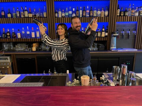 Hinter einer Bar stehen ein Mann und eine Frau Rücken an Rücken und lächeln in die Kamera. Sie halten beide Cocktailshaker in den Händen.