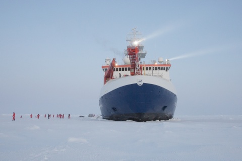 Ein Forschungsschiff liegt auf Eis und Menschen in orangefarbenen Anzügen laufen daneben.