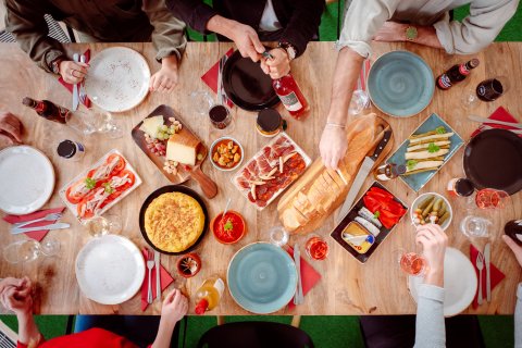 Ein gedeckter Tisch von oben, an dem sechs Menschen sitzen. Zu sehen sind verschiedene spanische Lebensmittel, wie Schinken, Käse und Wein. 